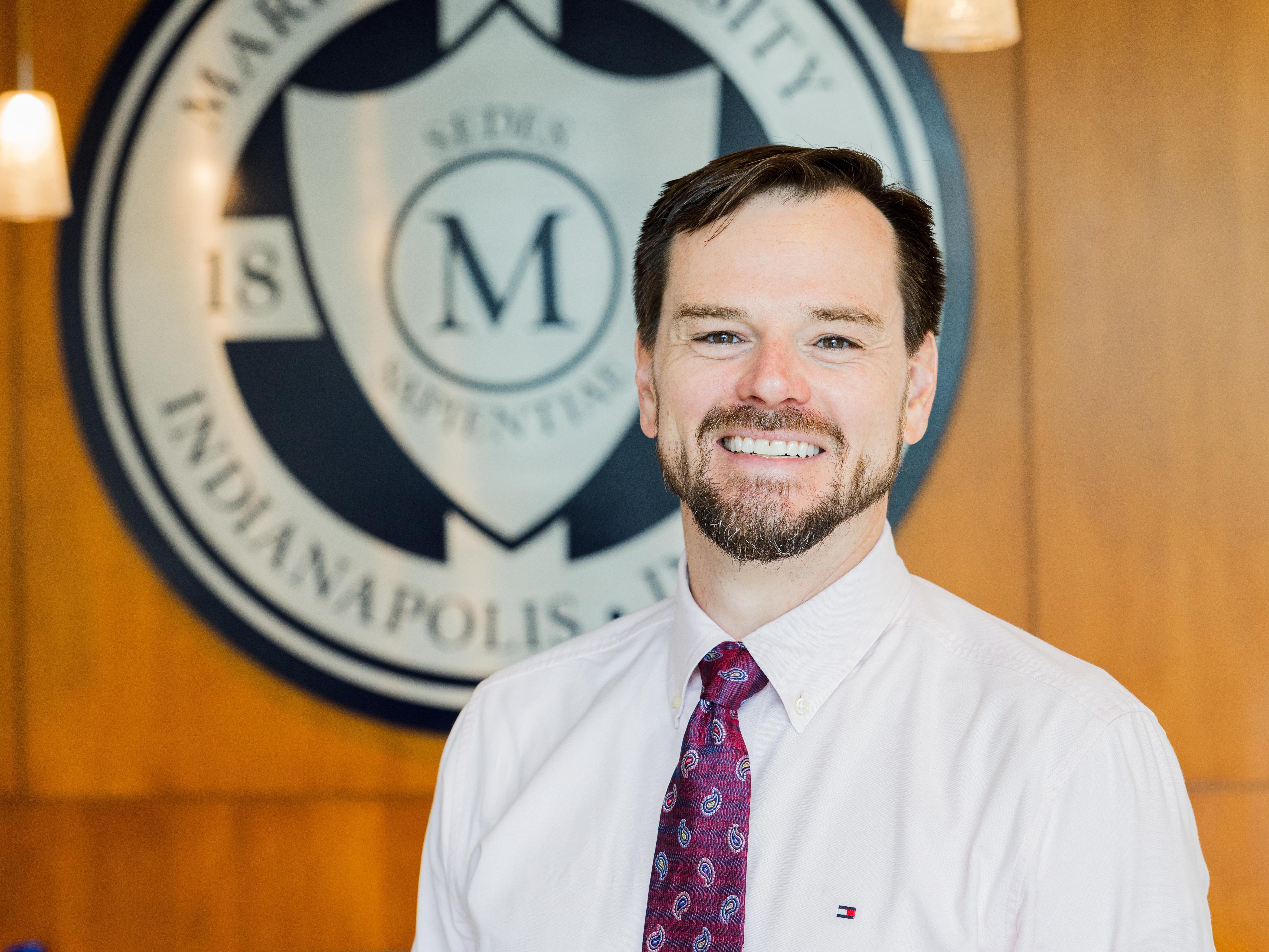 Smiling man in a dress shirt and tie standing indoors with a large circular emblem featuring a letter “M” blurred in the background.