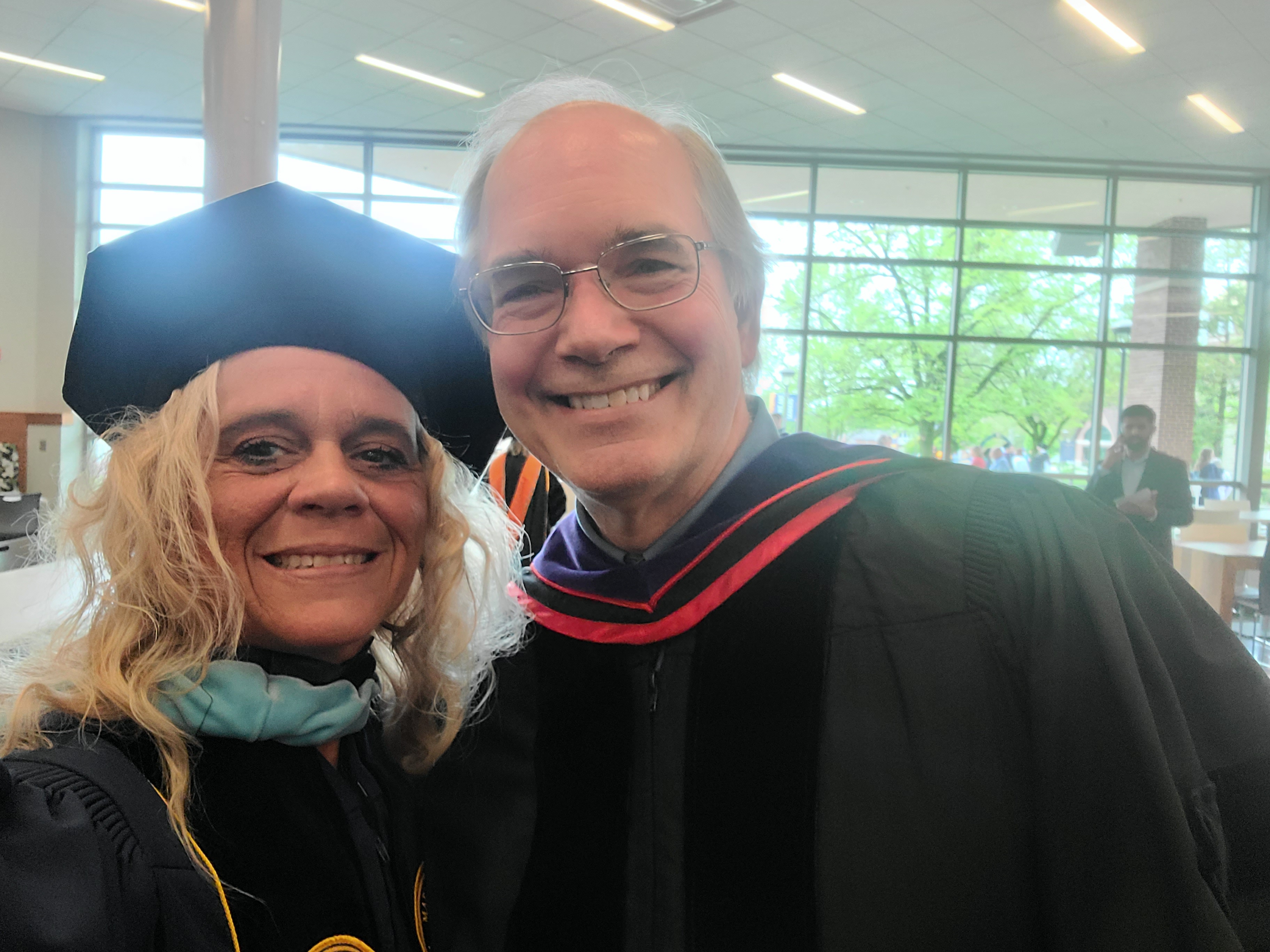 Two individuals in graduate garb posing for a selfie at a graduation ceremony, featuring a woman in a graduation cap and gown alongside a man in academic regalia.