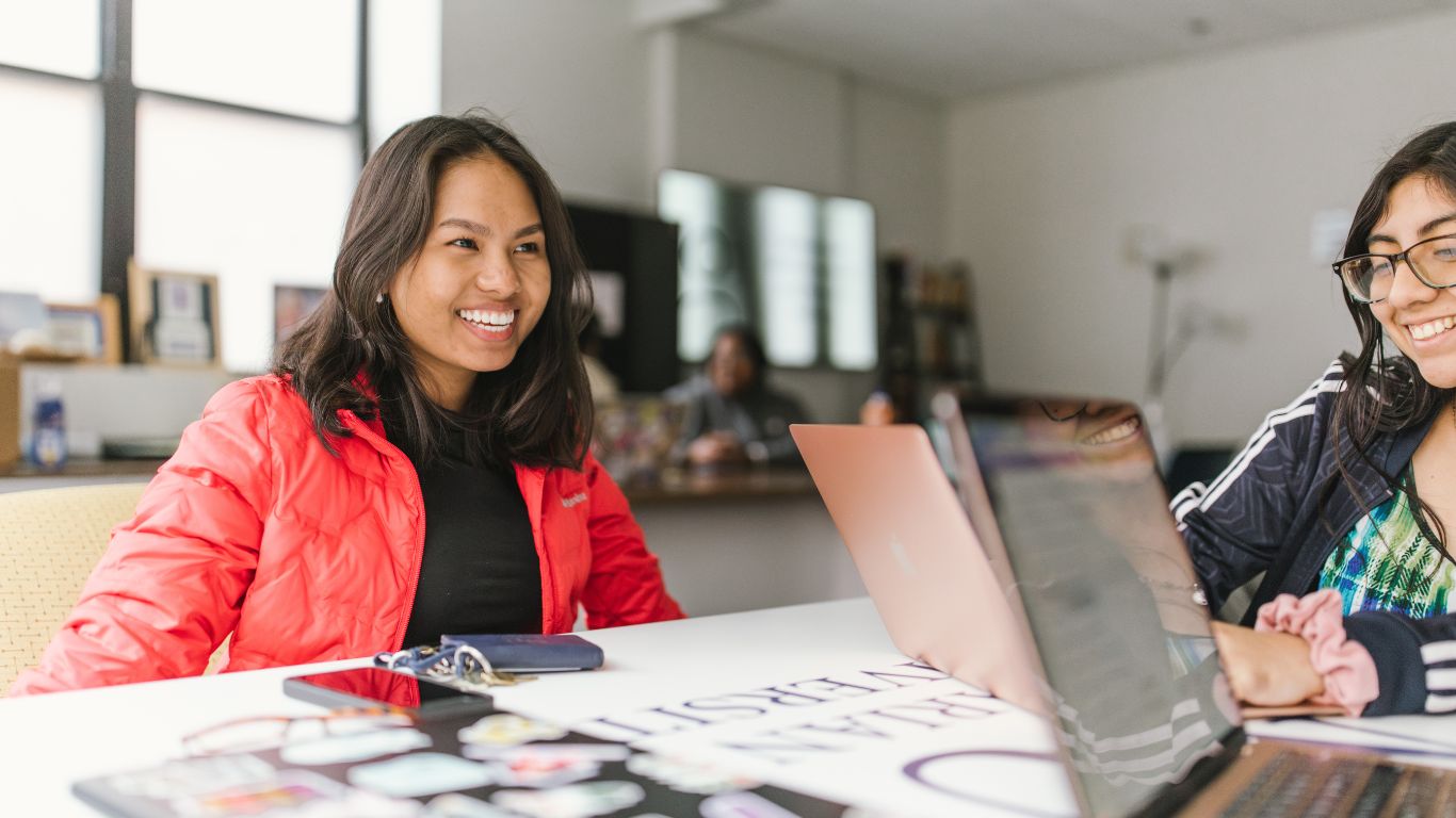Two women engaged in a conversation at a table with laptops, showcasing a collaborative and friendly environment.