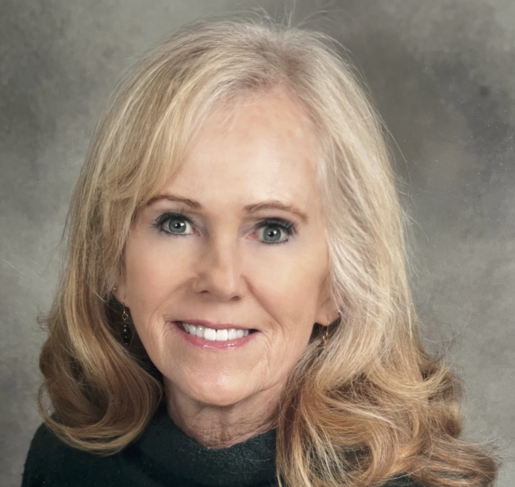 Smiling older woman with short blonde hair wearing a light blue blazer outdoors with trees in the background.