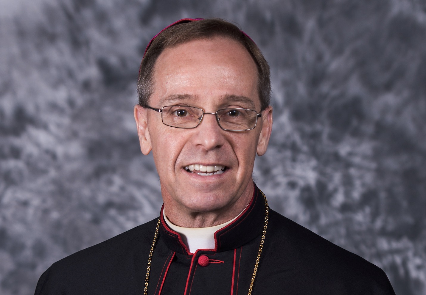 Smiling clergyman wearing glasses and a black cassock with red trim against a gray studio backdrop.