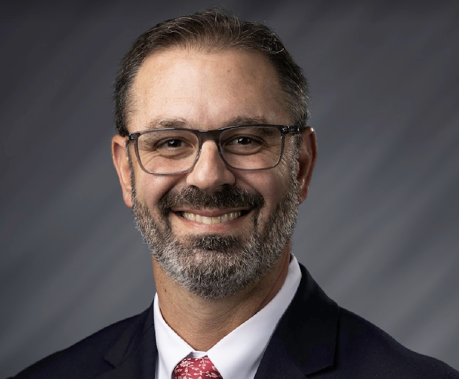 Smiling man in a suit and red tie wearing glasses, in a professional headshot against a gray background.