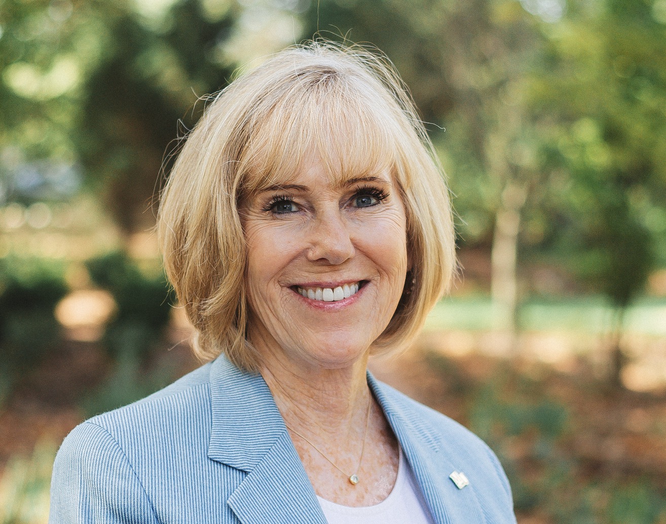 Smiling older woman with short blonde hair wearing a light blue blazer outdoors with trees in the background.