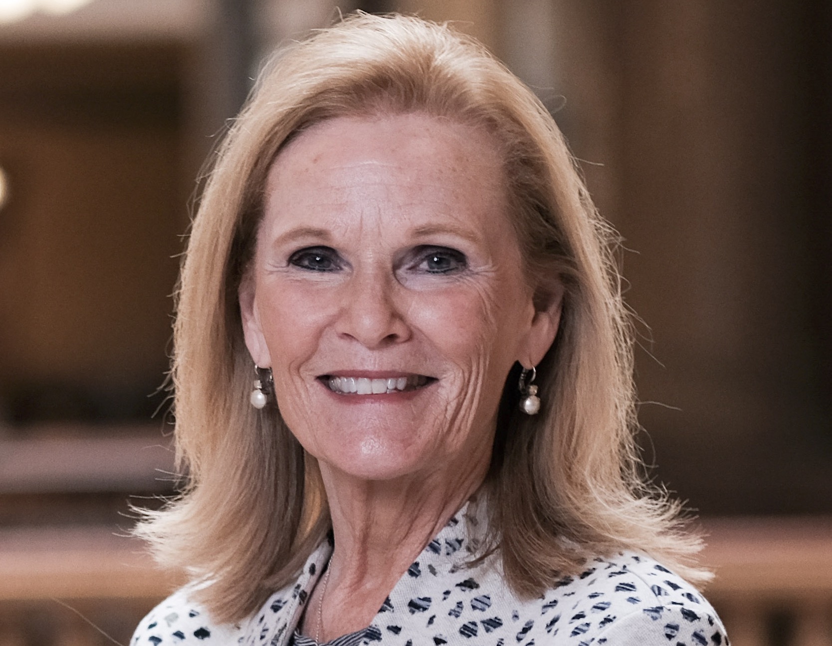 Smiling woman with blonde hair wearing pearl earrings, in a softly lit indoor setting.