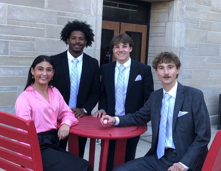 Four Marian University School of Business students dressed in professional attire sit and stand around a red outdoor table after winning the 2024 Indiana University National Team Selling Championship.