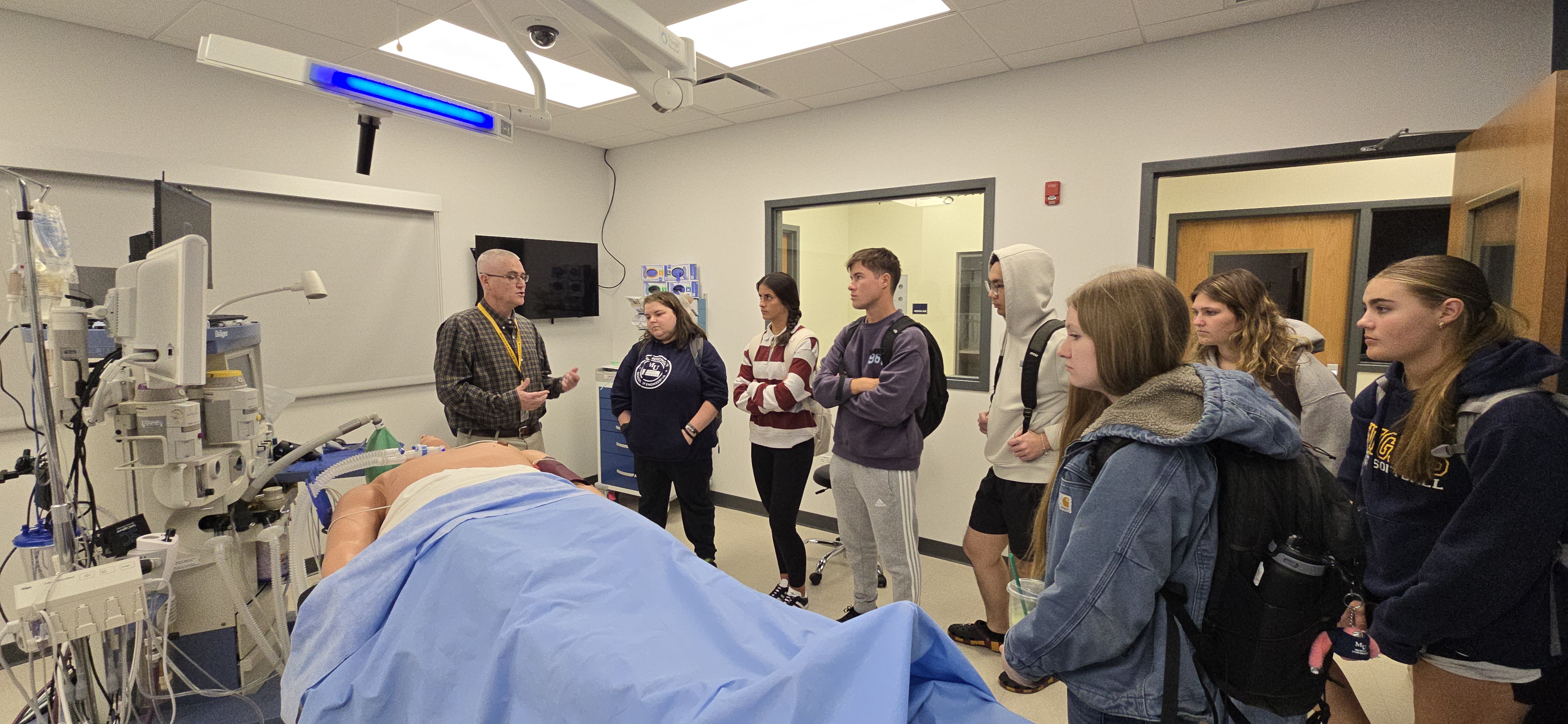 Instructor explains medical equipment beside a hospital bed in a simulation lab while a group of students listens.