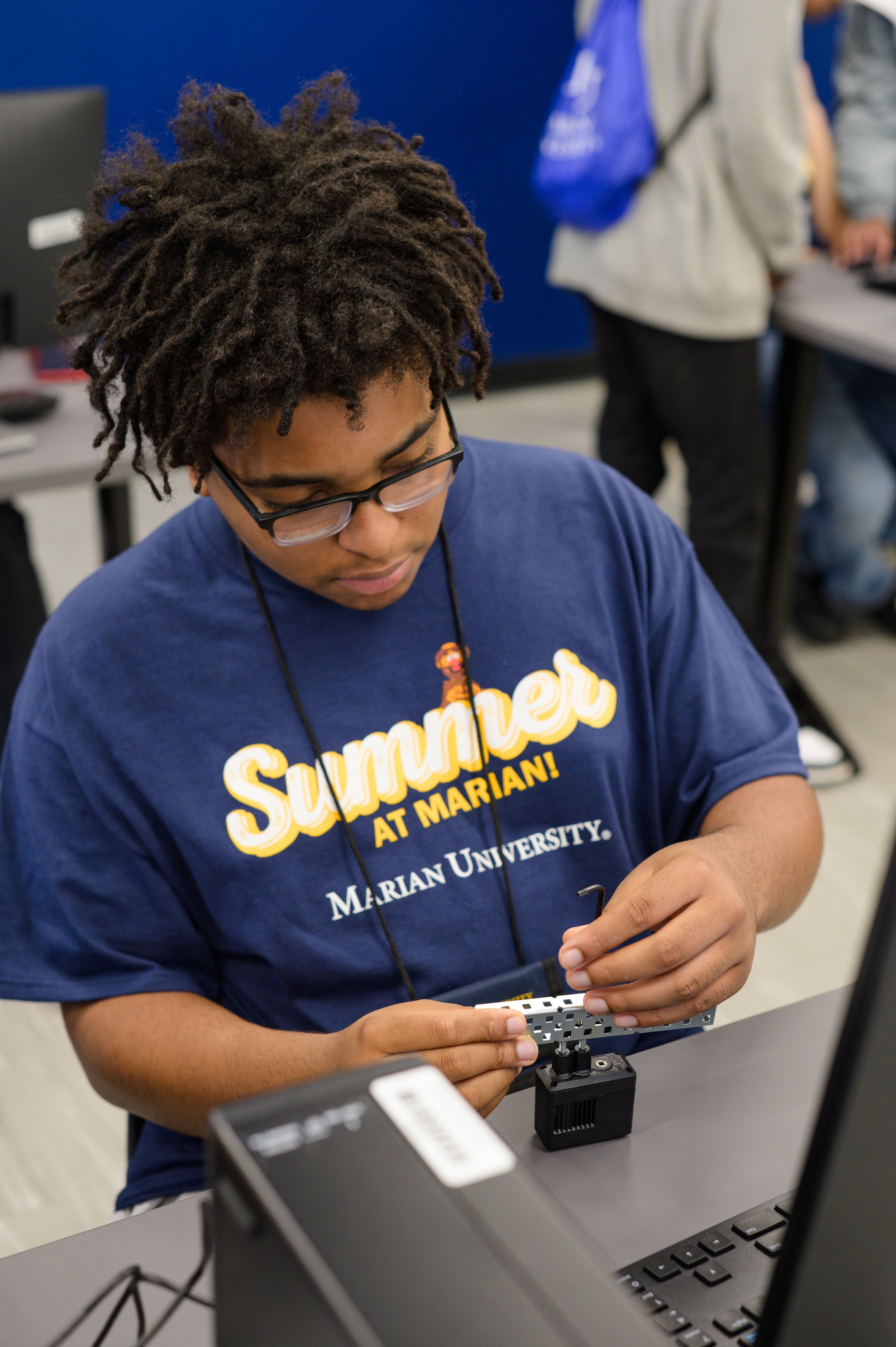 A student in a "Summer at Marian University" t-shirt works on assembling components at a computer workstation.